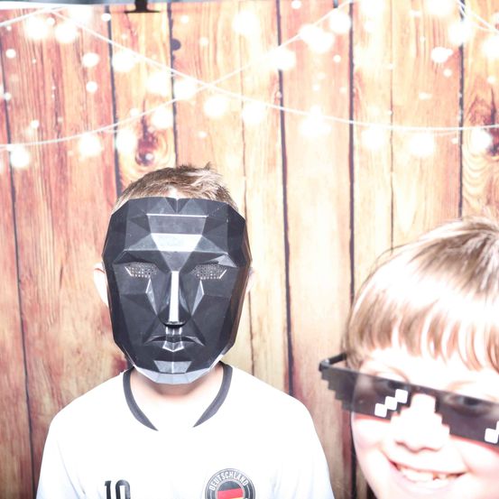 Zwei Jungen posieren für ein Foto. Einer trägt eine schwarze geometrische Maske und der andere hat Minecraft-Stil-Brillen. Hinter ihnen ist ein hölzerner Hintergrund mit Lichterketten.