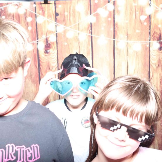 Drei Kinder posieren für ein Foto. Der Junge in der Mitte trägt eine Totenkopfhaube und herzförmige Brille. Das Mädchen rechts trägt pixelierte Brille. Sie lächeln vor einem hölzernen Hintergrund.