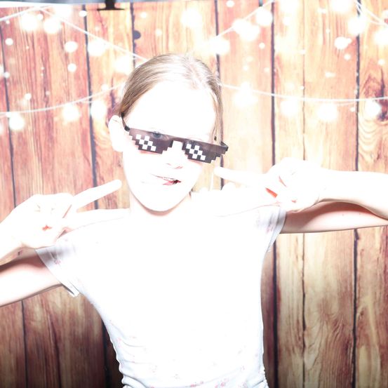 Ein junges Mädchen posiert für ein Foto mit pixeligen Brillengläsern und macht ein Friedenszeichen mit ihren Händen vor einem hölzernen Hintergrund.