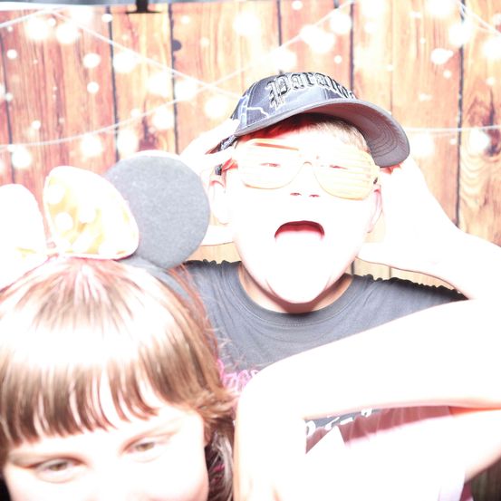 Ein junger Junge und ein Mädchen posieren für ein Foto in einem Fotokabine, wobei der Junge eine Sonnenbrille und eine Kappe trägt und das Mädchen Micky-Maus-Ohren trägt. Sie stehen vor einer Holzwand mit einer festlichen Dekoration.