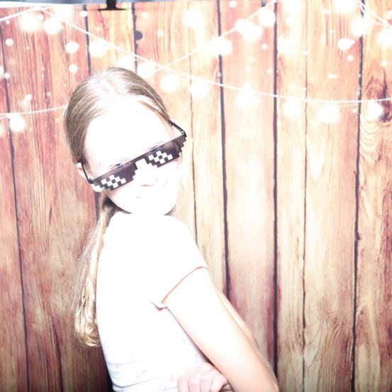 Ein junges Mädchen mit einer Brille mit pixeliertem Muster steht vor einer Holzwand mit Lichtern.