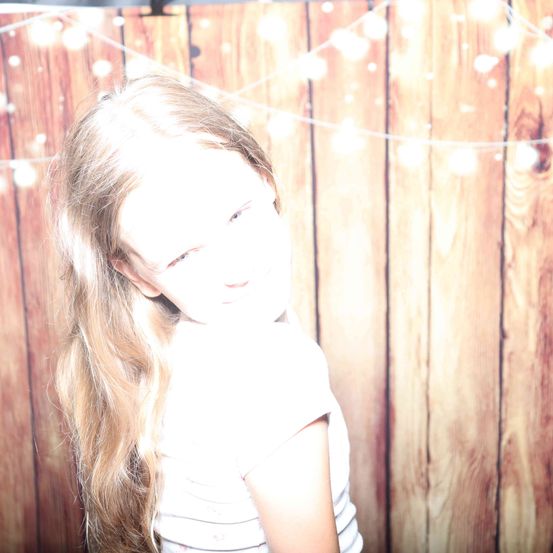 Ein Mädchen mit langen blonden Haaren in einem weißen T-Shirt steht vor einem hölzernen Hintergrund mit Lichterketten.
