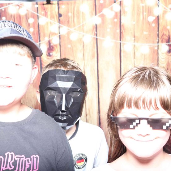 Drei Kinder posieren für ein Foto. Der Junge auf der linken Seite trägt eine Baseball-Kappe, der in der Mitte trägt eine Maske. Das Mädchen auf der rechten Seite trägt Pixel-Gläser. Sie stehen vor einer Holzwand.