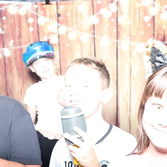 Ein Junge hält ein Mikrofon und lächelt. Ein Mädchen mit einer Katzenohren-Haarspange steht neben ihm. Ein weiterer Junge und ein Mädchen stehen hinter ihm. Sie stehen vor einer Holzwand mit Lichtern.