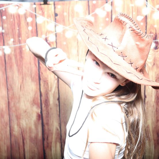 Ein junges Mädchen mit langen blonden Haaren, das einen braunen Cowboyhut und ein weißes T-Shirt trägt, posiert für ein Foto vor einem hölzernen Hintergrund.