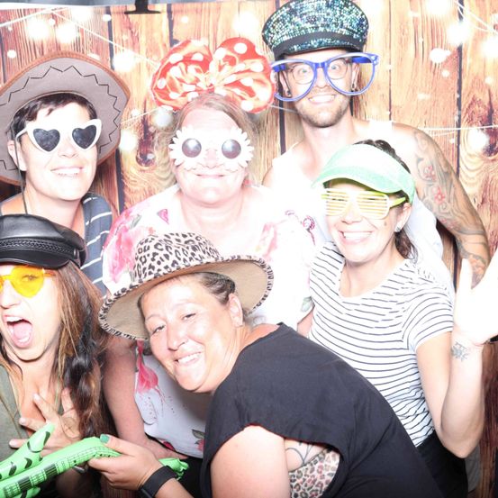 Eine Gruppe von Menschen in verschiedenen Hüten und Sonnenbrillen posiert für ein Foto in einem Fotoautomaten mit einem hölzernen Hintergrund.