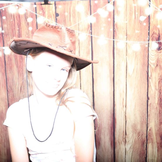 Ein junges Mädchen mit einem braunen Cowboyhut und einer Schnur um den Hals, vor einem holzverkleideten Hintergrund mit Lichtern.