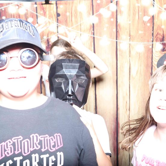 Ein junger Mann mit einer zerrissenen grauen T-Shirt und einer Sonnenbrille posiert mit einem Mädchen und einer Person mit Maske hinter ihm in einer Fotokabine.