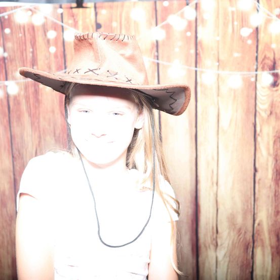 Ein Mädchen in einem Cowboyhut mit weißem Shirt und einer Halskette posiert vor einem hölzernen Hintergrund mit Lichterketten.