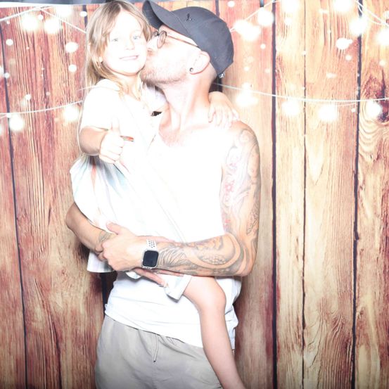 Ein Mann mit Tätowierungen küsst ein junges Mädchen auf die Wange vor einer Holzwand mit Lichterketten.