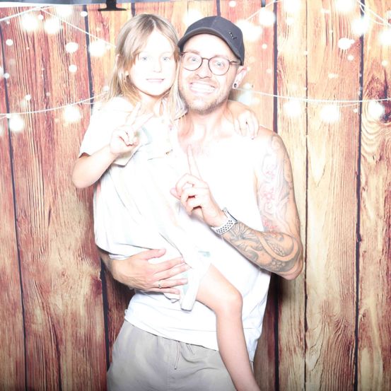 Ein Mann und ein junges Mädchen stehen vor einer Holzwand. Der Mann trägt eine Mütze und eine Brille, das Mädchen ein weißes Hemd. Beide lächeln und posieren für ein Foto.