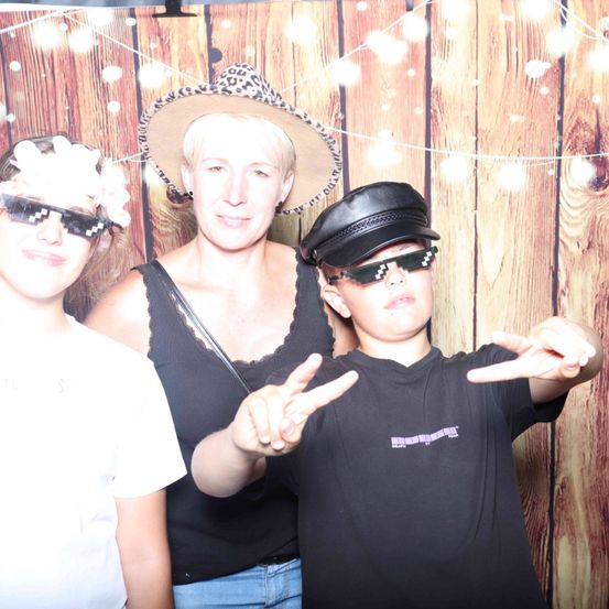 Drei Personen posieren für ein Foto vor einem hölzernen Hintergrund. Die Frau trägt einen Leopardenhut und der Junge trägt eine schwarze Mütze und eine Sonnenbrille. Das Mädchen trägt ein weißes T-Shirt und eine Sonnenbrille.