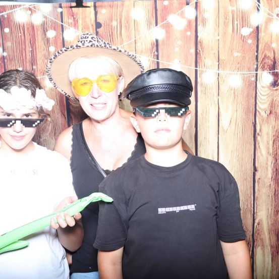 Eine Frau und zwei Kinder posieren für ein Foto vor einem hölzernen Hintergrund. Die Frau trägt eine Mütze und eine Sonnenbrille, und die Kinder tragen auch Sonnenbrillen und halten eine grüne Spielfigur.