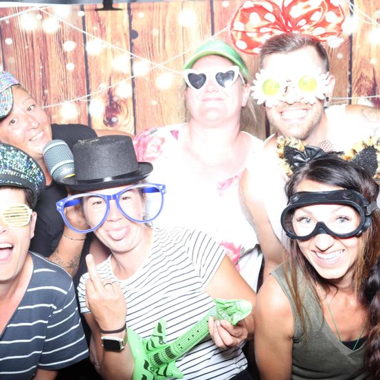 Eine Gruppe von Menschen posiert für ein Foto in einem Fotokabinen mit hölzernem Hintergrund, trägt verschiedene Hüte und Brillen. Eine Frau hält eine grüne Gitarre.