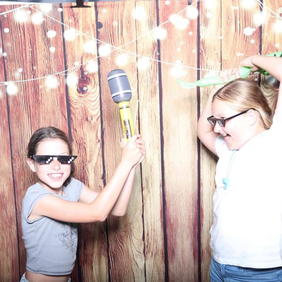 Zwei Mädchen posieren für ein Foto in einem Fotokabinen-Hintergrund. Eine hält ein Mikrofon, die andere ein grünes Spielzeug. Beide tragen Sonnenbrillen.