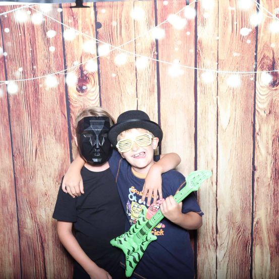 Zwei junge Jungen posieren für ein Foto vor einem hölzernen Hintergrund. Ein Junge trägt eine schwarze Maske, während der andere einen schwarzen Hut trägt und eine grüne Spielzeuggitarre hält.