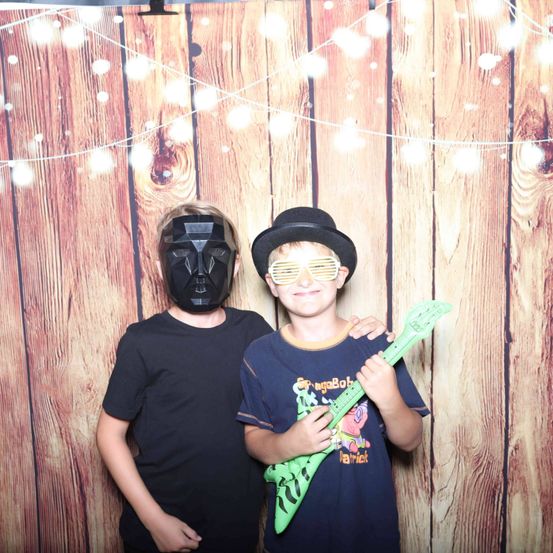 Zwei junge Jungen stehen vor einem hölzernen Hintergrund. Ein Junge trägt eine Darth Vader-Maske, während der andere eine grüne Gitarre hält.