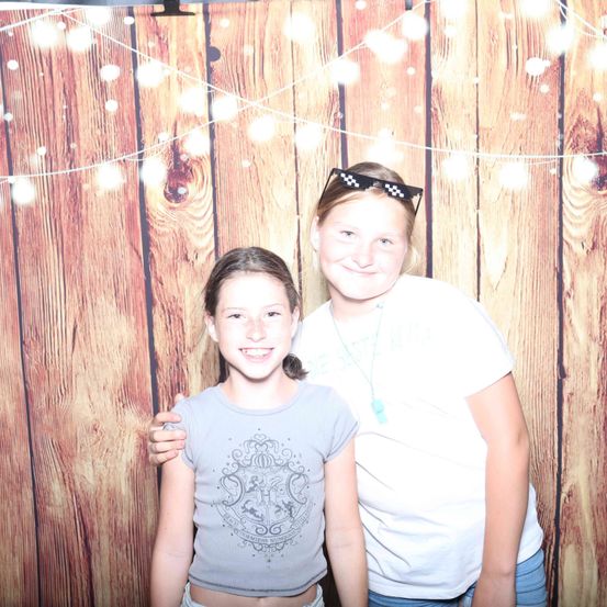 Zwei Mädchen stehen eng beieinander und posieren für ein Foto vor einem hölzernen Hintergrund mit Lichterketten.