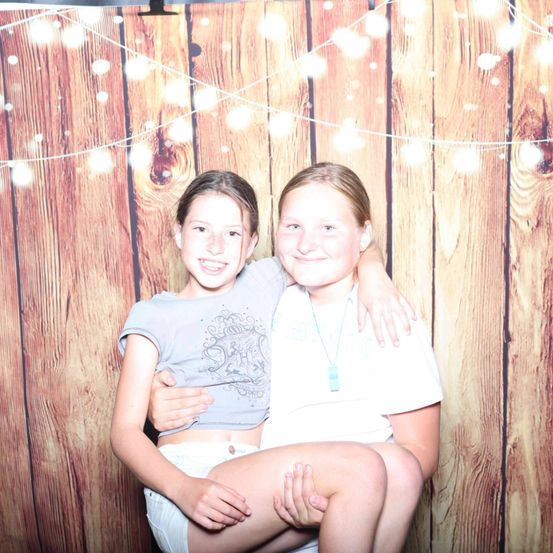 Zwei junge Mädchen sitzen eng beieinander, lächeln und posieren für ein Foto vor einer Holzwand mit Lichterketten.
