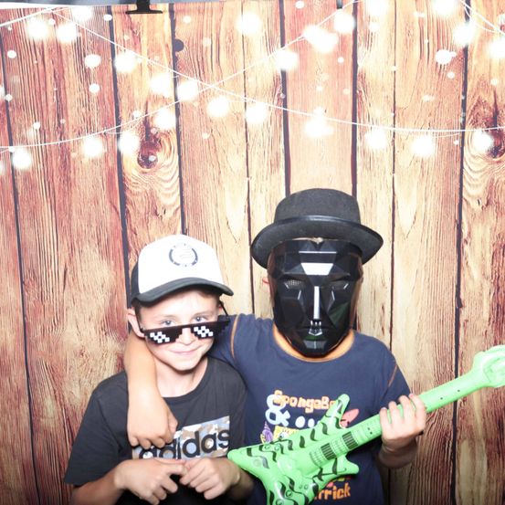 Zwei junge Jungen stehen vor einer hölzernen Kulisse mit Lichterketten. Einer trägt eine schwarze Maske und hält eine grüne Gitarre, während der andere eine weiße Kappe und eine Brille trägt.