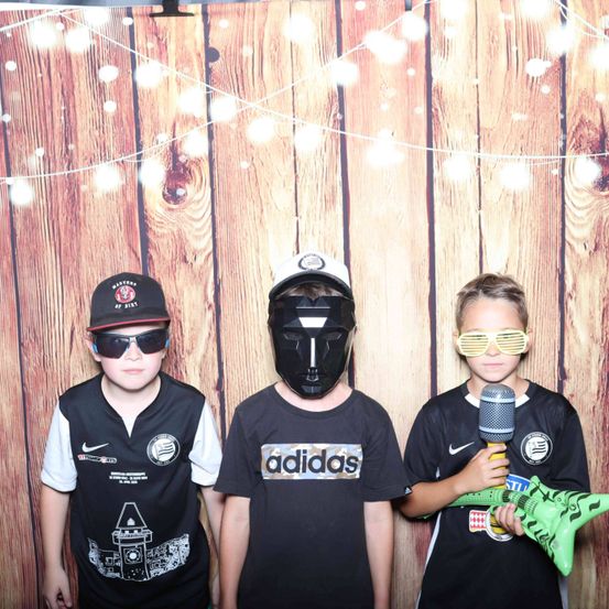 Drei Jungen stehen zusammen in einem Fotokabinen. Der Junge auf der linken Seite trägt eine Baseballkappe und Sonnenbrille, der Junge in der Mitte hat eine schwarze Maske und der Junge auf der rechten Seite hält ein Mikrofon und eine grüne Gitarre. Sie tragen alle schwarze Hemden.