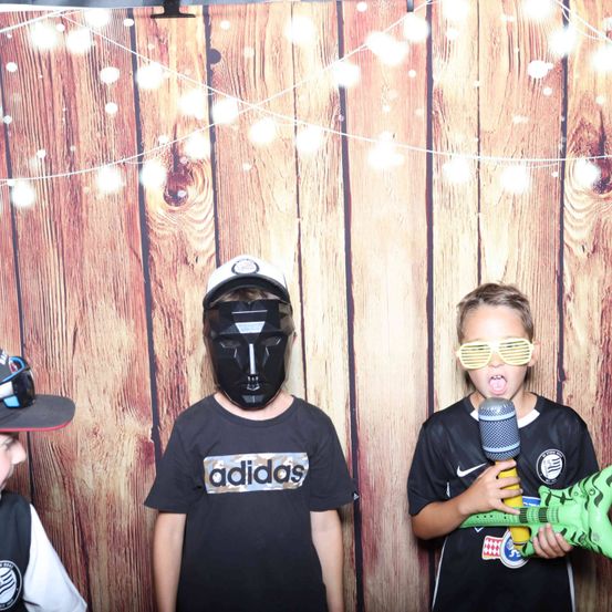 Drei Jungen vor einem hölzernen Hintergrund mit Lichtern. Der Junge auf der linken Seite trägt einen Hut, der Junge in der Mitte trägt eine Maske und der Junge auf der rechten Seite hält ein Spielzeugmikrofon.