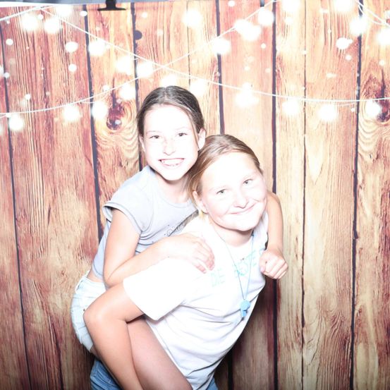 Zwei junge Mädchen stehen eng beieinander und lächeln vor einem hölzernen Hintergrund mit Lichterketten.
