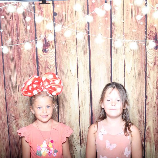 Zwei junge Mädchen stehen nebeneinander und posieren für ein Foto. Das Mädchen auf der linken Seite trägt ein rosa Shirt mit einem roten Bogen auf dem Kopf. Das Mädchen auf der rechten Seite trägt ein rosa Tanktop mit Schmetterlingsaufdruck. Dahinter befindet sich ein hölzerner Hintergrund mit Lichterketten.