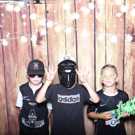 Drei junge Jungen stehen vor einem hölzernen Hintergrund mit Lichtern. Der Junge links trägt einen schwarzen Hut mit weißen Buchstaben. Der Junge in der Mitte trägt eine schwarze Maske und macht ein Friedenszeichen. Der Junge rechts hält eine grüne Spielzeuggitarre.