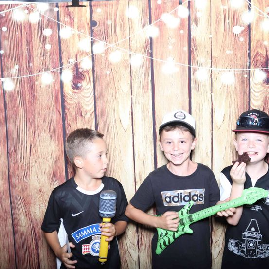 Drei Jungen stehen vor einer Holzwand. Der Junge auf der linken Seite hält ein Mikrofon. Der Junge in der Mitte hält eine Gitarre. Der Junge auf der rechten Seite hält ein Stück Kuchen.