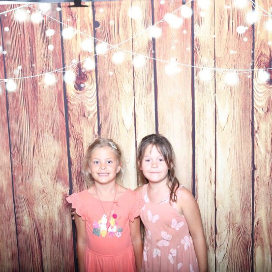 Zwei junge Mädchen stehen vor einem hölzernen Hintergrund mit Lichterketten. Beide Mädchen tragen rosa Kleider.
