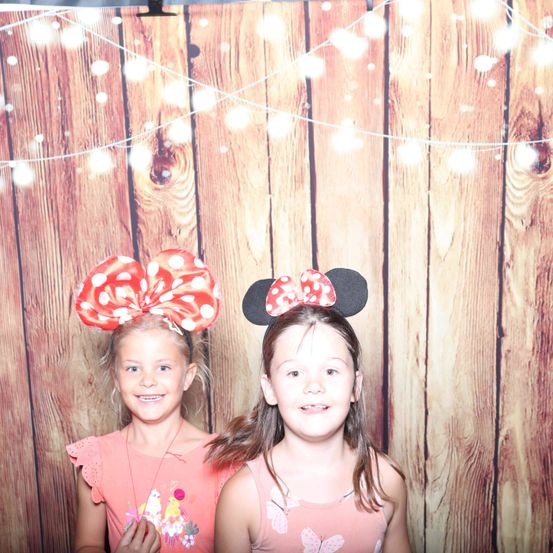 Zwei junge Mädchen, beide lächelnd, stehen eng beieinander in einem Fotokabine. Sie tragen Minnie-Mouse-Ohren. Hinter ihnen ist eine Holzwand mit Lichterketten.