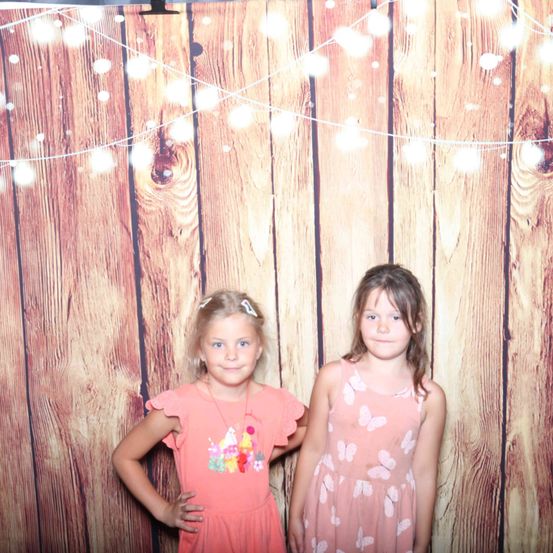 Zwei junge Mädchen stehen nebeneinander vor einer Holzwand mit Lichterketten. Beide Mädchen tragen rosa Kleider. Das Mädchen auf der linken Seite hat ein Kuchendesign auf ihrem Kleid.