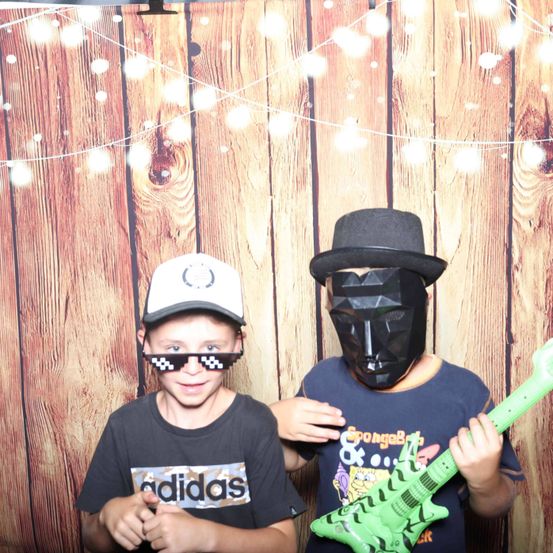 Zwei Jungen posieren für ein Foto. Einer trägt einen Hut und eine Sonnenbrille, der andere ein Maske und hält eine grüne Gitarre. Sie stehen vor einer Holzwand mit Lichterketten.