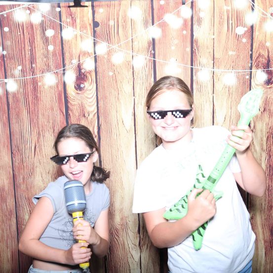 Zwei Mädchen in einem Fotostudio mit einem hölzernen Hintergrund, eine hält ein Mikrofon und die andere eine grüne Gitarre. Sie tragen beide Pixelbrillen.