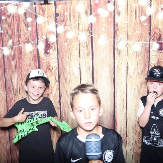 Drei Jungen stehen vor einem hölzernen Hintergrund. Der Junge auf der linken Seite hält eine grüne Spielzeuggitarre. Der Junge in der Mitte hält ein Mikrofon und der Junge auf der rechten Seite isst etwas.