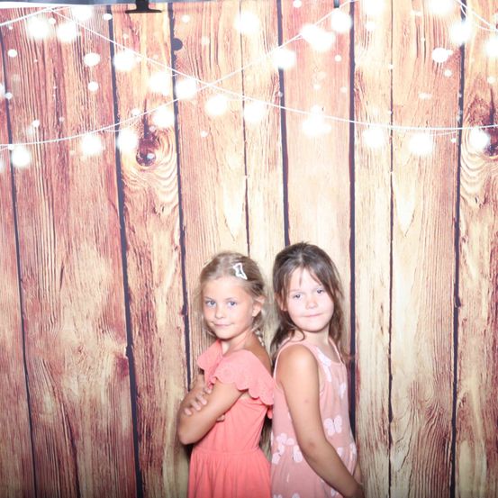 Zwei junge Mädchen stehen vor einer Holzwand mit Lichterketten. Das Mädchen auf der linken Seite hat die Arme verschränkt und das Mädchen auf der rechten Seite hat die Hände hinter dem Rücken. Sie schauen beide in die Kamera.
