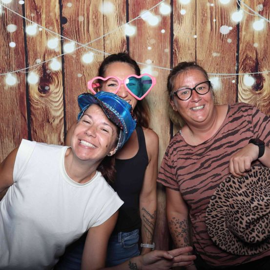 Drei Frauen posieren für ein Foto vor einem hölzernen Hintergrund mit Lichterketten. Die Frau auf der linken Seite trägt ein weißes T-Shirt und einen Hut. Die mittlere Frau hat herzförmige Brillen. Die Frau auf der rechten Seite trägt ein braunes Shirt mit Zebramuster und hält einen Leoparden-Print-Hut.