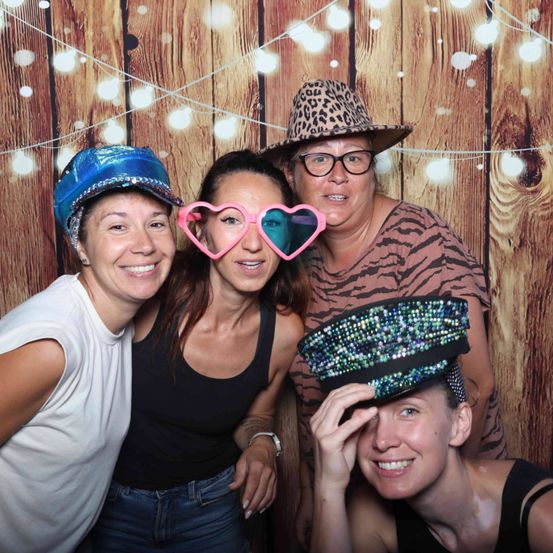 Vier Frauen lächeln und posieren für ein Foto. Sie tragen Hüte und Sonnenbrillen. Eine Frau trägt herzförmige Brillen. Sie stehen vor einer Holzwand mit Lichtern.