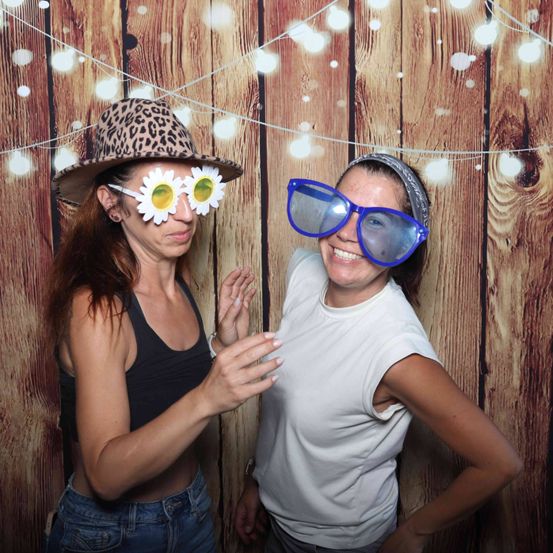 Zwei Frauen vor einem hölzernen Hintergrund mit weißen Lichtern. Eine trägt einen Leopardenmusterhut und Sonnenblumen-Sonnenbrille, die andere ein weißes T-Shirt mit blauer Brille.