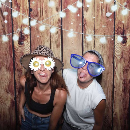 Zwei Frauen posieren vor einer Holzwand und tragen lustige Brillen. Eine trägt einen Leopardenmusterhut mit blumenförmigen Gläsern, die andere eine übergroße blaue Brille.
