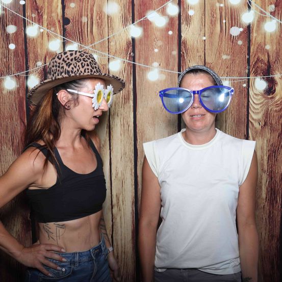 Zwei Frauen stehen vor einem hölzernen Hintergrund. Die Frau auf der linken Seite trägt einen Leopardenmusterhut mit Gänseblümchengläsern und einem schwarzen Tanktop. Die Frau auf der rechten Seite trägt blaue Sonnenbrille und ein weißes ärmelloses Oberteil.