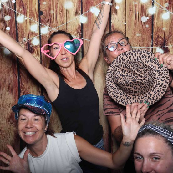 Vier Frauen posieren für ein Foto in einem Studio mit einem hölzernen Hintergrund. Eine trägt herzförmige Brillen, eine andere einen Leopardenmusterhut, während zwei weitere lächeln und winken.
