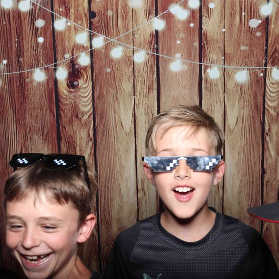 Zwei Jungen stehen vor einem hölzernen Hintergrund mit Lichterketten. Der Junge auf der linken Seite trägt eine Sonnenbrille auf dem Kopf, während der Junge auf der rechten Seite pixelierte Brille trägt.