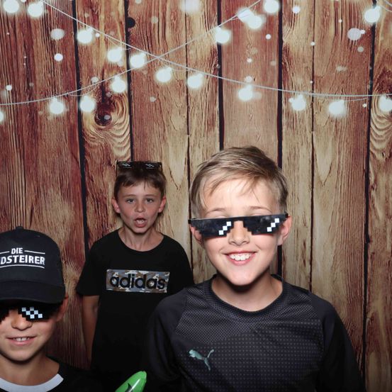 Drei Jungen stehen vor einer Holzwand mit Lichterketten. Der Junge in der Mitte trägt ein schwarzes T-Shirt mit einem weißen Adidas-Logo und einer schwarzen Mütze. Der Junge auf der rechten Seite trägt eine Sonnenbrille und ein schwarzes T-Shirt mit einem weißen Schachbrettmuster. Der Junge auf der linken Seite trägt eine schwarze Kappe mit dem Wort 'DESTEIRER'.