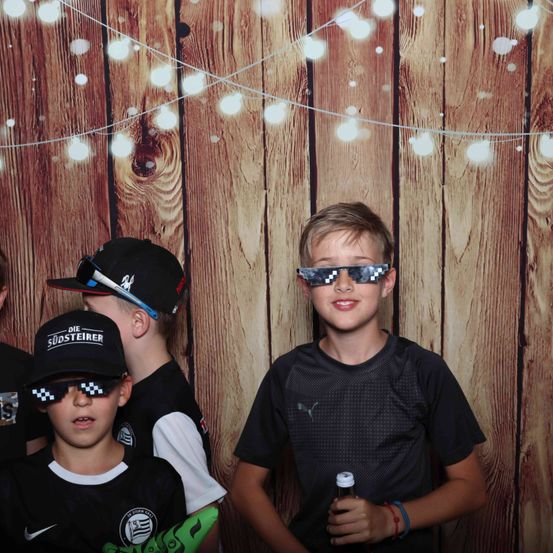 Drei Jungen posieren vor einem hölzernen Hintergrund mit Lichterketten. Sie tragen schwarze Shirts, Mützen und Sonnenbrillen. Einer hält eine Flasche.