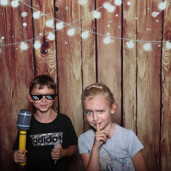Zwei Kinder, ein Junge und ein Mädchen, stehen vor einem hölzernen Hintergrund mit Lichterketten. Der Junge hält ein Mikrofon und trägt eine Sonnenbrille, während das Mädchen ihren Finger an die Lippen hält.