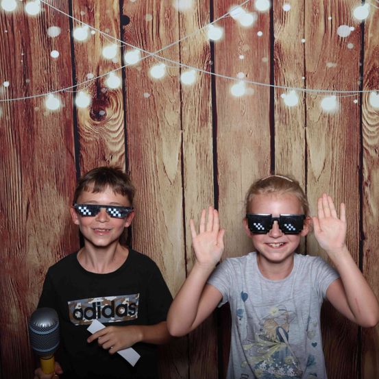 Zwei Kinder stehen vor einer Holzwand mit Lichterketten, lächeln und posieren für ein Foto. Ein Kind hält ein Mikrofon und beide tragen Sonnenbrillen.