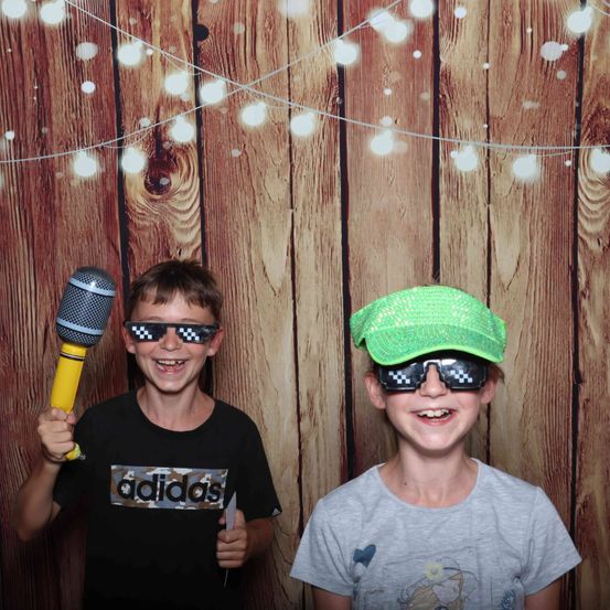 Zwei junge Jungen posieren für ein Foto vor einem hölzernen Hintergrund. Einer hält ein Mikrofon, der andere trägt einen grünen Hut. Beide tragen Sonnenbrillen.