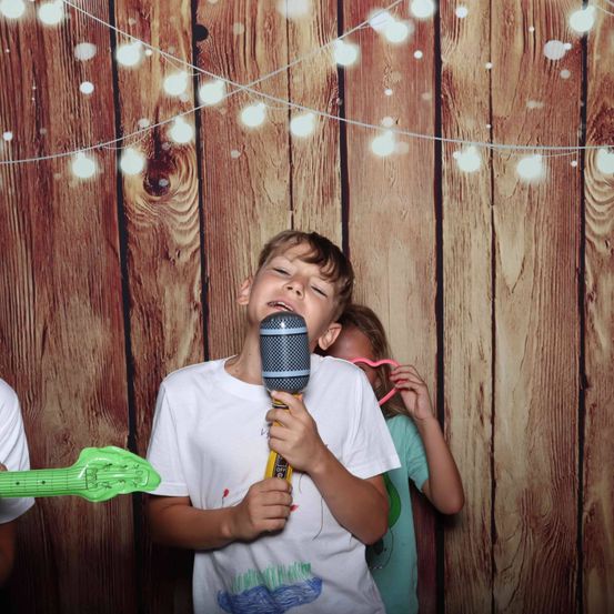 Ein junger Junge hält ein Spielzeugmikrofon und singt in einem Fotostudio, während ein Mädchen mit rosa Brille zusieht. Die Holzwand ist mit weißen Lichtern dekoriert.